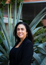 Maria L Orozco headshot, dark medium length hair, wearing a black blouse, standing in front of an agave plant