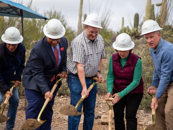 individuals in a row, holding shovels and breaking ground in desert scene