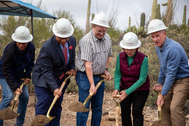 individuals in a row, holding shovels and breaking ground in desert scene