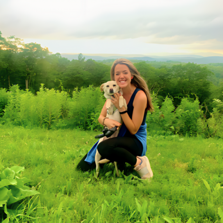 Alison Irwin kneeling in a grassy field, holding a small dog, sunset in the distance