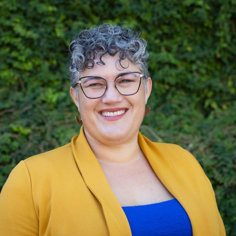 heather wearing yellow jacket, blue blouse, glasses and salt and pepper hair, greenery in background